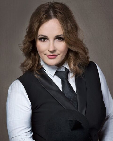 Portret satyryczki i aktorki Joanny Kołaczkowskiej. Ma pofalowane, brązowe włosy i jest ubrana w elegancki strój w stylu power dressing: białą koszulę, czarną kamizelkę i czarny krawat w delikatny wzór. Patrzy prosto w obiektyw z pewnym siebie wyrazem twarzy, trzymając w ręku (częściowo widoczny) czarny kapelusz. Tło jest ciepło-szare.