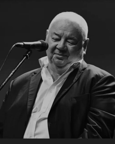 Czarno-białe ujęcie polskiego muzyka i wokalisty Stanisława Soyki podczas występu. Artysta siedzi przy mikrofonie, z zamkniętymi oczami i lekkim, skupionym uśmiechem, pogrążony w śpiewie. Ma krótkie, jasne włosy. Jest ubrany w ciemną marynarkę i białą koszulę z rozpiętym kołnierzykiem.