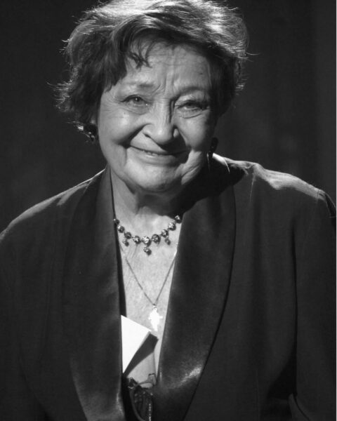 Czarno-białe zbliżenie na aktorkę Ewę Dałkowską. Uśmiecha się, patrząc prosto w obiektyw. Ma krótkie, ciemne włosy i jest ubrana w ciemny strój, prawdopodobnie marynarkę ze szpiczastymi, satynowymi klapami. Nosi naszyjnik z zawieszką w kształcie krzyża.