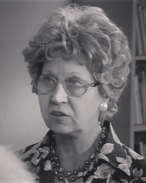 Czarno-białe zbliżenie na aktorkę Mirosławę Marcheluk. Ma krótkie, kręcone włosy i nosi duże okulary w jasnej oprawce. Ubrana jest w bluzkę lub sukienkę we wzór kwiatowy i ma na sobie naszyjnik z dużych, okrągłych koralików oraz perłowe kolczyki. Patrzy z intensywnym wyrazem twarzy, prawdopodobnie podczas sceny filmowej lub teatralnej. Tło jest rozmyte, z widocznym regałem na książki.