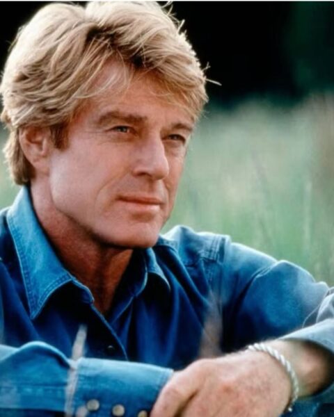 Portret Roberta Redforda w średnim wieku, aktora o jasnych włosach, ubranego w niebieską koszulę dżinsową. Spogląda w bok, na jego nadgarstku widać srebrną bransoletkę. W tle delikatnie rozmyta zieleń natury. Zdjęcie oddaje klasyczną urodę i charyzmę gwiazdora, idealnie wpisując się w inspirujące treści o kulturze.