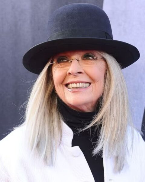 Zbliżenie na amerykańską aktorkę Diane Keaton na czerwonym dywanie. Uśmiecha się szeroko i ma na sobie charakterystyczny dla niej strój: czarny kapelusz z szerokim rondem, okrągłe okulary w cienkiej oprawce z bursztynowymi szkłami, czarny golf oraz białą, pikowaną kurtkę lub marynarkę. Ma długie, siwe włosy. Jest to ikona mody, której styl jest inspiracją dla kobiet z klasą ceniących oryginalność i elegancję.
