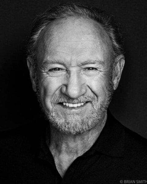 Czarno-biały, portret aktora Gene'a Hackmana. Uśmiecha się szeroko, patrząc prosto w obiektyw. Ma krótkie, siwe włosy, jasną, przystrzyżoną brodę i jest ubrany w ciemną koszulkę polo. Zdjęcie ma studyjny charakter z ciemnym tłem i podkreśla charakter oraz charyzmę tej hollywoodzkiej legendy, idealnie pasującej do artykułów o kulturze i inspirujących ikonach kina.