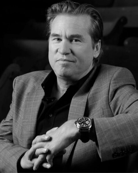 Czarno-biały portret aktora Vala Kilmera. Siedzi w jasnej, wzorzystej marynarce i czarnej koszuli, spoglądając prosto w obiektyw z poważnym wyrazem twarzy. Na lewym nadgarstku ma duży, ciemny zegarek na pasku. Ręce ma splecione, opierając się na oparciu krzesła lub ławki. Tło jest ciemne i rozmyte, co podkreśla elegancję i dojrzały styl aktora – inspiracja dla treści o kulturze i męskiej modzie z klasą.