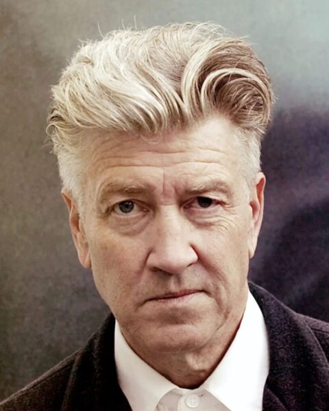 Zbliżenie na twarz reżysera Davida Lyncha. Ma surowy, skupiony wyraz twarzy i charakterystyczną fryzurę: zaczesane do góry, siwe włosy z jasnymi pasemkami. Jest ubrany w białą koszulę i ciemną marynarkę. Zdjęcie ma studyjny charakter z nieostrym, ciemniejszym tłem.