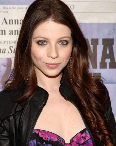 Zbliżenie na aktorkę Michelle Trachtenberg. Ma długie, kasztanowe włosy, jasną cerę i intensywnie niebieskie oczy, podkreślone ciemnym makijażem. Jest ubrana w czarną, skórzaną kurtkę i gorset w fioletowo-różowy, kwiatowy wzór. Spogląda prosto w obiektyw z delikatnym uśmiechem.