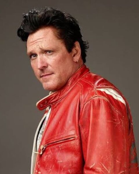 Portret aktora Michaela Madsena, ujęcie z profilu, na którym patrzy przez ramię prosto w obiektyw. Ma ciemne, zaczesane do góry włosy i kolczyk w uchu. Jest ubrany w czerwoną, skórzaną kurtkę z białymi akcentami. Tło jest neutralne, szare.