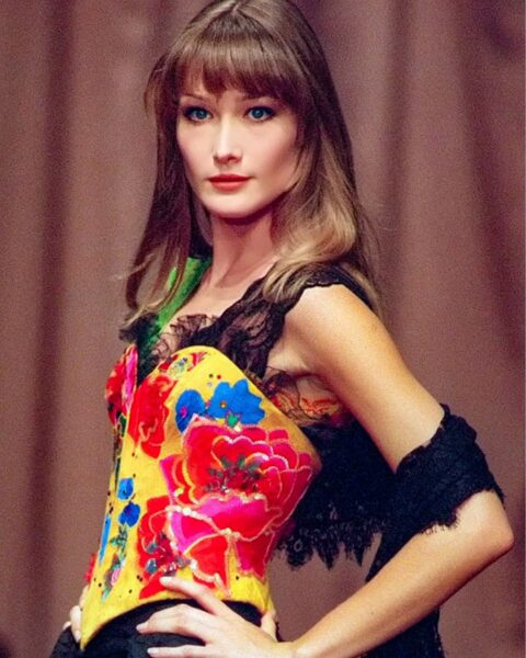 Carla Bruni: Zjawiskowe ujęcie top modelki lat 90. w trakcie pokazu, prezentującej elegancką, czarną kreację. Jej charakterystyczny, uwodzicielski wzrok i klasyczna uroda były kluczowe dla zdefiniowania luksusowej estetyki tej supermodelkowej dekady.