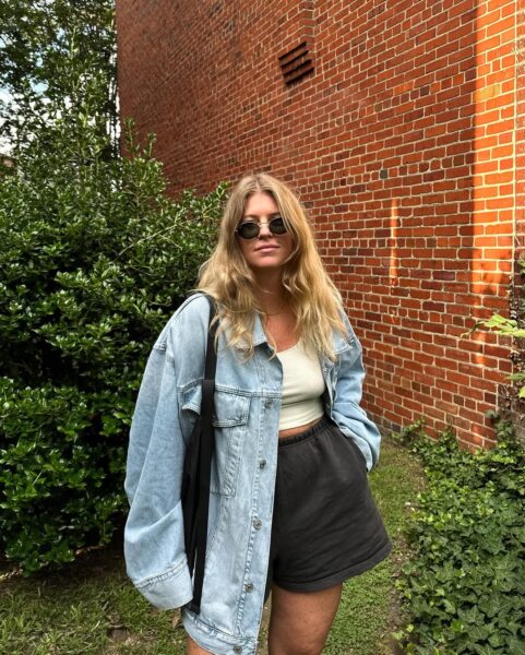 Kobieta o blond włosach, pozująca na zewnątrz przy ceglanym murze i zielonej roślinności. Ma na sobie kluczowy, ponadczasowy element garderoby: luźną, oversizową kurtkę dżinsową (w stylu "shacket" lub "boyfriend"), którą narzuciła na prosty, kremowy top na ramiączkach. Dół stanowią luźne, ciemne szorty, a całość uzupełniają klasyczne, okrągłe okulary przeciwsłoneczne. Stylizacja jest kwintesencją casualowego komfortu i mody, która nie przemija.