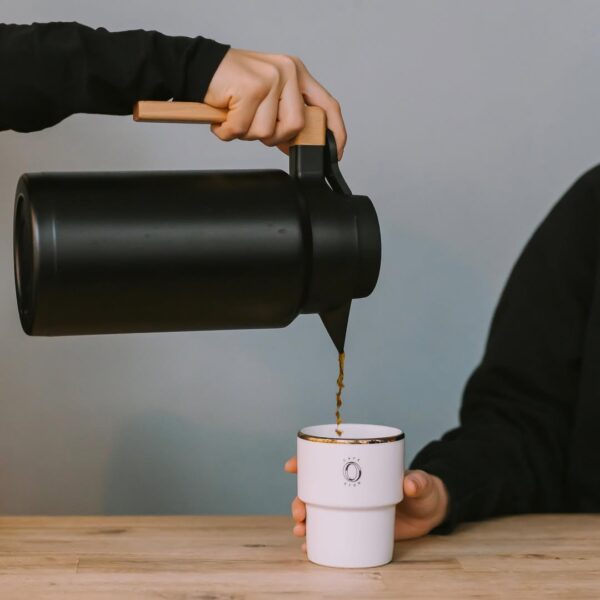 Zbliżenie na minimalistyczny moment parzenia kawy, gdzie dłoń nalewa ciemny napój z czarnego, skandynawskiego termosu z drewnianą rączką do białej, prostej filiżanki. Scena ta doskonale wpisuje się w klimat skandynawskich kawiarni w Warszawie, akcentując prostotę i wysoką jakość podania kawy na drewnianym stole.