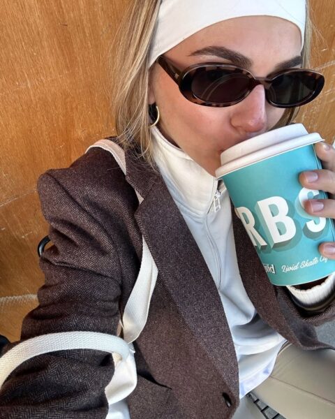 Ujęcie selfie z bliska, gdzie kobieta pije kawę z kubka w kolorze turkusowym. Ma na sobie białą opaskę na włosach, która jest prostym i modnym rozwiązaniem, idealnym jako szybka fryzura dla leniwych. Stylizację dopełniają okulary przeciwsłoneczne i brązowa marynarka.