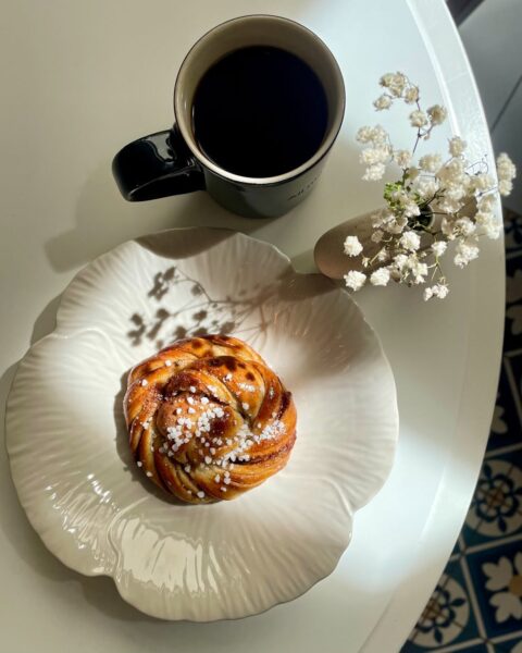 Jasny i ciepły kadr z góry przedstawia klasyczną szwedzką bułeczkę cynamonową (kanelbullar) posypaną grubym cukrem perlistym, umieszczoną na białym, fantazyjnym talerzyku w kształcie kwiatu. Obok na białym stoliku stoi czarna filiżanka z kawą oraz miniaturowy wazonik z delikatną gipsówką, całość skąpana w naturalnym świetle i klimacie skandynawskiej kawiarni.
