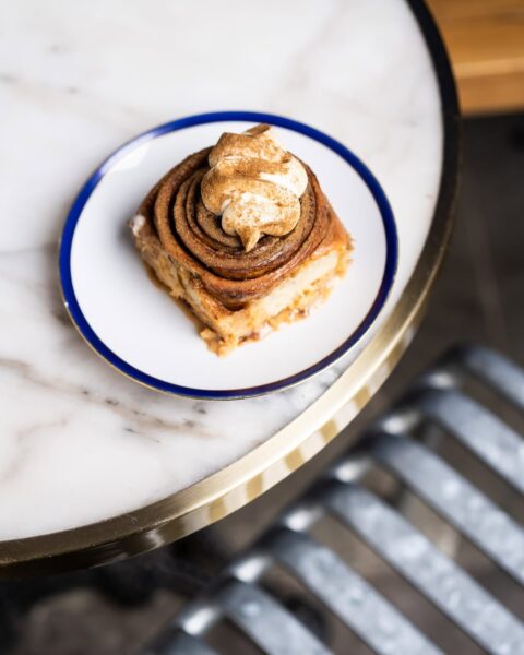 Zbliżenie z góry na bogato glazurowaną bułeczkę cynamonową, zwieńczoną bitą śmietaną i cynamonem, serwowaną na małym talerzyku z granatowo-złotym rantem. W tle widać elegancki stolik z blatem z białego marmuru i złotą obwódką, co świadczy o wysokiej estetyce, jaką cenią skandynawskie kawiarnie w Warszawie.