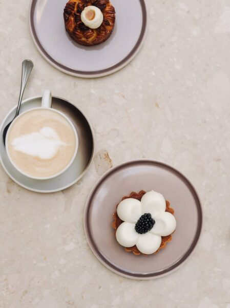Ujęcie z góry przedstawia trzy eleganckie desery i filiżankę kremowego latte ze wzorem na spienionym mleku, ustawione na jasnym, marmurowym blacie. Dwa ciastka w kształcie kwiatów, w tym jedno z jeżyną, serwowane na pastelowych talerzykach, podkreślają dbałość o detale i wysoką jakość słodkości, z których słyną najlepsze skandynawskie kawiarnie w Warszawie.