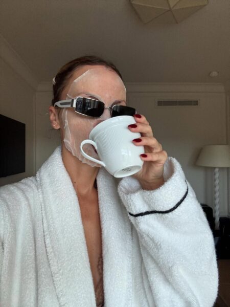 Zdjęcie przedstawia kobietę w białym, puszystym szlafroku, która oddaje się porannym rytuałom pielęgnacyjnym, pijąc kawę z białego kubka. Na jej twarzy widać nawilżającą maseczkę w płachcie, a na oczach spoczywają stylowe okulary przeciwsłoneczne, dodające nuty nonszalancji. Cała scena podkreśla wysiłek włożony w codzienną dbałość o cerę suchą mimo nawilżania, szukając ukojenia i intensywnej regeneracji. Zdjęcie to wizualizuje moment relaksu i odnowy, dążąc do poprawy kondycji skóry po nocy.