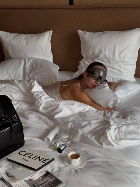 Kobieta leży wygodnie w łóżku pod białą pościelą z opaską do spania na oczach (lub maską pielęgnacyjną na czoło) oraz okularami. Obok niej leżą kosmetyki, magazyny i filiżanka kawy.