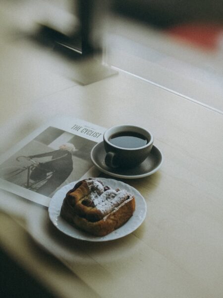 Kadr utrzymany w minimalistycznym i stonowanym stylu, przedstawiający ciemną kawę w szarej filiżance, świeżą drożdżówkę posypaną cukrem pudrem oraz fragment magazynu "The Cyclist" na jasnym drewnianym parapecie. Ta prosta, estetyczna kompozycja doskonale oddaje nastrój i atmosferę, którą oferują skandynawskie kawiarnie w Warszawie.