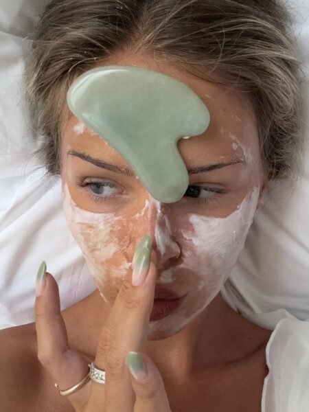 Zdjęcie prezentuje kobietę w trakcie relaksującej i intensywnej pielęgnacji, wykonując masaż twarzy płytką gua sha z jadeitu. Na jej skórze widoczna jest gruba, biała maseczka, która jest częścią zaawansowanego rytuału mającego przynieść ulgę. Ujęcie to symbolizuje poszukiwanie skutecznych metod na suchą skórę mimo nawilżania i stosowania wielu kosmetyków. Tego typu domowe spa ma na celu głęboką regenerację i odżywienie cery, aby wreszcie przełamać chroniczne uczucie ściągnięcia.