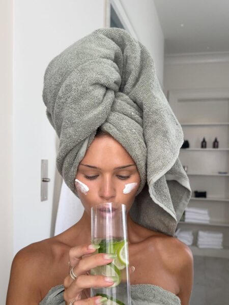 Zdjęcie ukazuje kobietę po kąpieli, owiniętą dużym, szarym ręcznikiem, która dba o nawodnienie organizmu, pijąc wodę z limonką. Na jej policzkach widoczne są dwie precyzyjnie nałożone porcje kremu do twarzy, co podkreśla ciągły nacisk na pielęgnację. Ujęcie to ilustruje wysiłek włożony w walkę z suchą skórą mimo nawilżania, zarówno zewnętrznego, jak i wewnętrznego. Cały kontekst zdjęcia sugeruje potrzebę kompleksowego podejścia do rutyny, aby faktycznie zregenerować i wzmocnić barierę hydrolipidową naskórka.