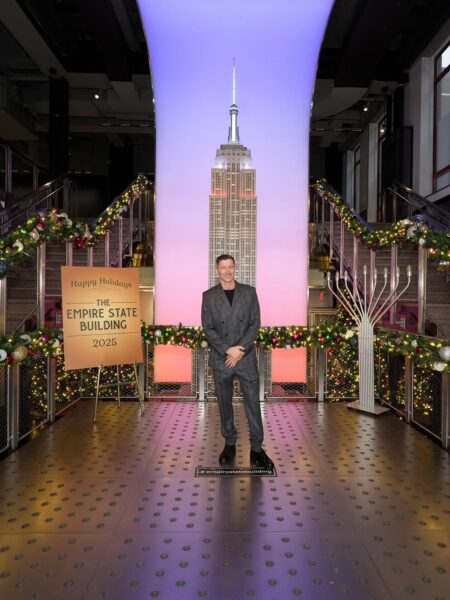 Robert Lewandowski w holu Empire State Building: Na pierwszym zdjęciu widzimy Roberta Lewandowskiego, ubranego w elegancki, szary garnitur w kratę, który stoi w holu Empire State Building. W tle za nim znajduje się podświetlona na fioletowo-różowo ściana z wizerunkiem wieżowca i świąteczną dekoracją. Jest to jedno z ujęć z historycznej wizyty, podczas której Lewandowscy na Empire State Building wzięli udział w uroczystości rozświetlenia budynku na biało-czerwono.