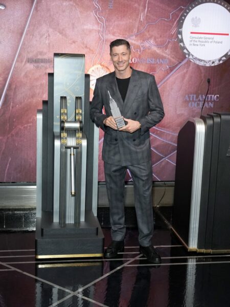 Robert Lewandowski, ubrany w szary garnitur, stoi przy art-decowskiej dekoracji, trzymając w dłoni miniaturową statuetkę Empire State Building. W tle widoczna jest duża mapa rejonu Nowego Jorku z logiem Konsulatu Generalnego RP w Nowym Jorku, co podkreśla oficjalny charakter wydarzenia, w którym uczestniczył.