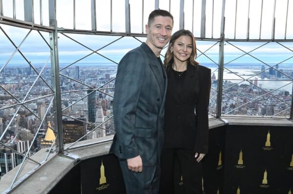 Zdjęcie przedstawia Lewandowskich na tarasie widokowym Empire State Building, podobnie jak na poprzednim ujęciu. Oboje uśmiechnięci i ubrani w ciemne, eleganckie stroje, pozują na tle rozległej panoramy Nowego Jorku. Jest to pamiątkowe zdjęcie z ich pobytu na szczycie wieżowca z okazji podświetlenia budynku na polskie barwy.