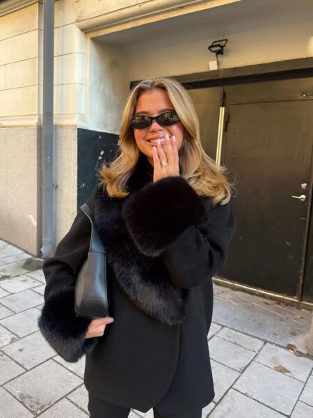 Elegancka blondynka w czarnym płaszczu z futrzanymi mankietami i kołnierzem uśmiecha się na ulicy, zasłaniając część ust dłonią. Jej stylowy, jesienny strój kontrastuje z jej rozświetloną twarzą, a błyszczący pierścionek na palcu przyciąga wzrok do jej uśmiechu. Mimo, że dłoń subtelnie zakrywa usta, widać jej jasne uzębienie, które wymaga szczególnej troski. Ten uśmiech przypomina, że nawet w pogoni za idealną sylwetką i modnym wyglądem, zdrowie jamy ustnej jest priorytetem, szczególnie w obliczu ryzyka powstawania ozempicowych zębów.