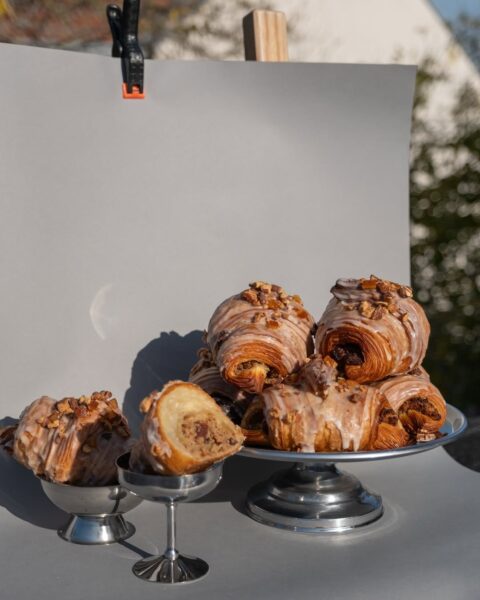 Kompozycja fotograficzna z rogalikami na dwóch metalowych paterach – większej i mniejszej – ustawionych na tle szarego papieru. Widać przekrój jednego z wypieków, ujawniający nadzienie z orzechów i bakalii. Ujęcie utrzymane w estetyce food photography, z miękkim światłem i surowym tłem.