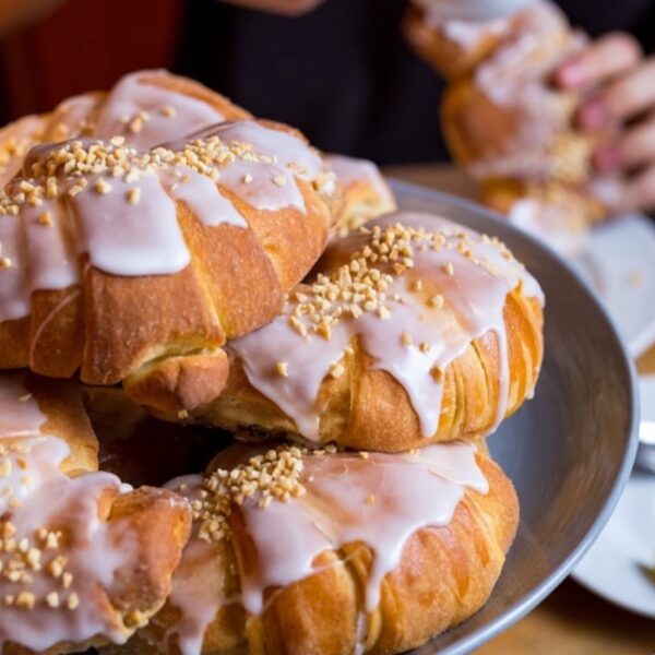 Talerz wypełniony świeżo upieczonymi rogalami polanymi lukrem i posypanymi drobno siekanymi orzechami. Ujęcie wykonane z bliska, w ciepłym świetle, które podkreśla złoty kolor ciasta i puszystą strukturę wypieków. W tle widoczna osoba trzymająca jeden z rogalików.