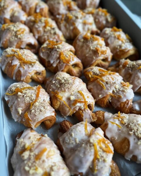 Jeden rogalik na beżowym talerzu, polany białym lukrem i posypany płatkami migdałów. Zdjęcie wykonane w miękkim świetle dziennym, z rozmytym tłem w jesiennych tonacjach, tworzy spokojny, estetyczny kadr w stylu slow food.