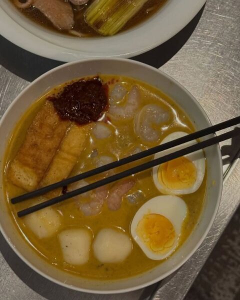 Zbliżenie na miskę aromatycznego ramenu w żółtym, kremowym bulionie, z dodatkiem krewetek, plastrów jajek na twardo z miękkim żółtkiem, chrupiącego tofu, pulpecików rybnych oraz porcji pikantnej pasty chili, z czarnymi pałeczkami opartymi o brzeg miski, sfotografowane na metalicznym blacie.