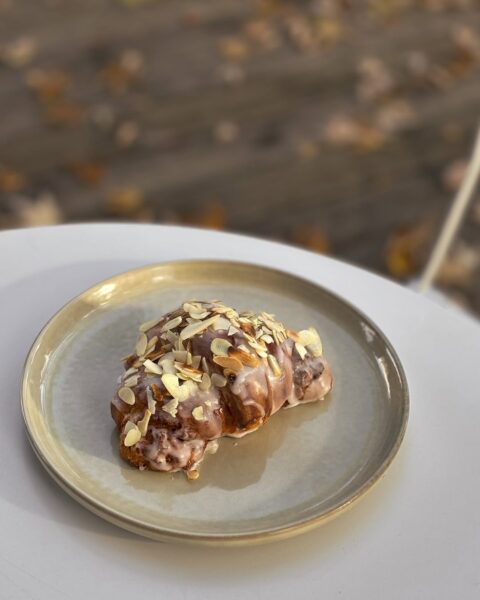 Zbliżenie na rogalik polany lukrem, udekorowany orzechami i trzymany przez dwie osoby — jedna dłoń podaje, druga odbiera wypiek na małym szklanym talerzyku. Minimalistyczna kompozycja w jasnym świetle podkreśla teksturę lukru i złocistą barwę ciasta.