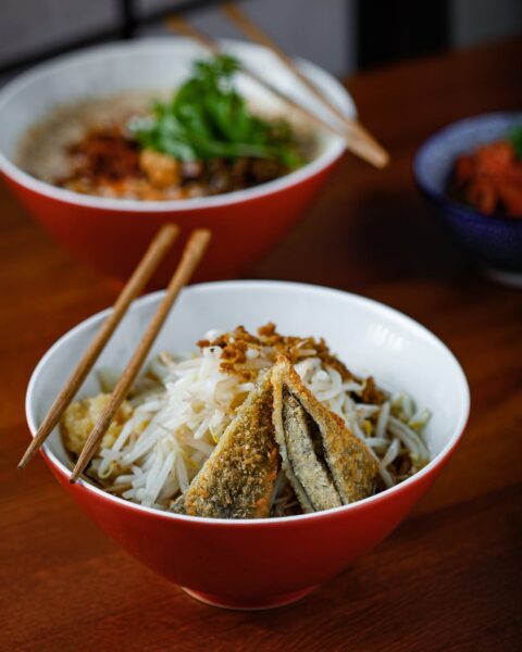 Biała miska z czerwonym zewnętrznym wykończeniem wypełniona makaronem, świeżymi kiełkami fasoli i chrupiącym smażonym filetem rybnym, ułożona na drewnianym stole, z drewnianymi pałeczkami opartymi o brzeg, w tle druga miska z ramenem i ziołami.