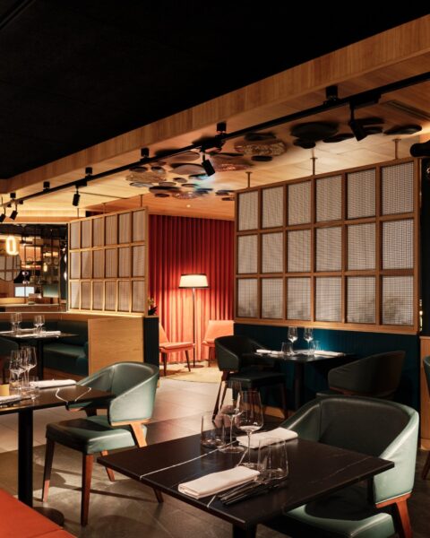 Stylowa restauracja o przyciemnionym, intymnym klimacie. Granatowe i zielone fotele zestawione z czarnymi stołami, białe serwetki i szkło ustawione z precyzją. Czerwone zasłony i lampy w tle wprowadzają teatralny akcent, a drewniane detale oraz siatkowe panele ścienne nadają wnętrzu japoński, współczesny charakter.