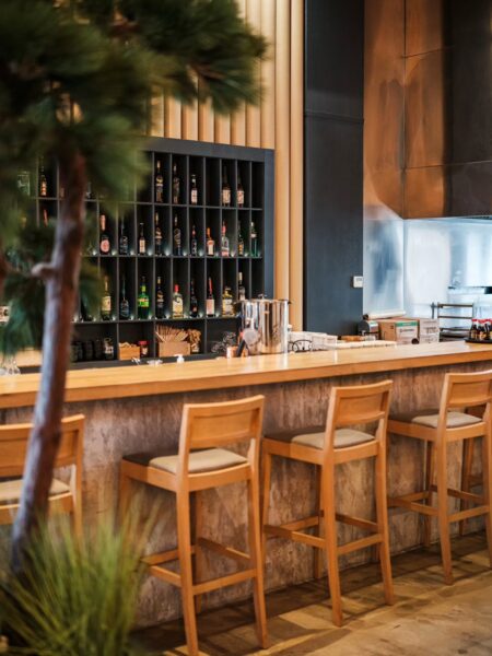Nowoczesny bar o wyraźnych japońskich inspiracjach – drewniany blat, wysokie krzesła w naturalnym kolorze i tło z geometryczną półką wypełnioną butelkami sake. Roślinność na pierwszym planie dodaje wnętrzu naturalności, a miękkie światło tworzy relaksującą, ciepłą atmosferę.