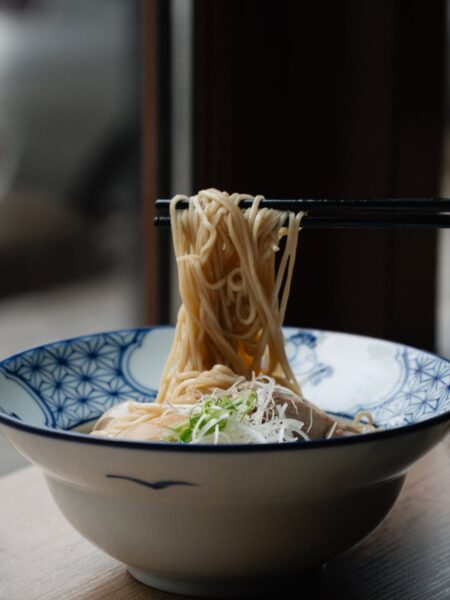 Pałeczki podnoszące porcję długiego, cienkiego makaronu ramen z pięknie zdobionej miski w niebiesko-białe wzory, z widocznymi dodatkami takimi jak cebulka i plaster mięsa, umieszczonej przy oknie z naturalnym światłem.