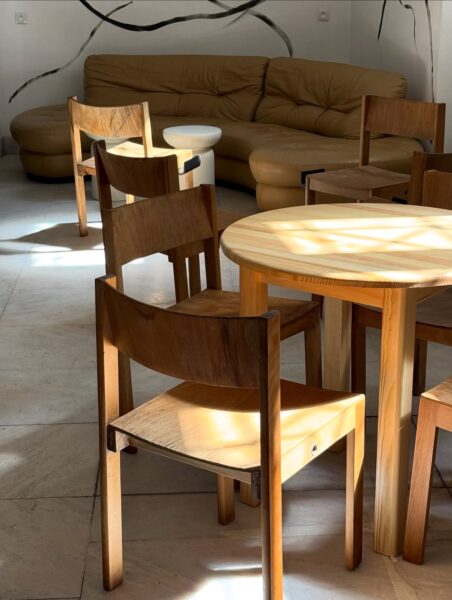 Wnętrze jasnej kawiarni z minimalistycznym wystrojem – drewniane krzesła i stoły w naturalnym kolorze, beżowa kanapa w tle i ściany ozdobione czarnymi, falującymi liniami. Promienie słońca tworzą świetliste refleksy na podłodze z jasnego kamienia.