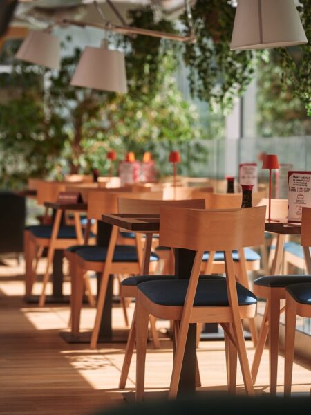 Jasne, przeszklone wnętrze restauracji z drewnianymi stołami i krzesłami o prostym, nowoczesnym designie. Słoneczne światło przenika przez roślinność w tle, tworząc przytulny, naturalny klimat. Na stołach stoją małe czerwone lampki i menu, nadające przestrzeni ciepły, gościnny charakter.