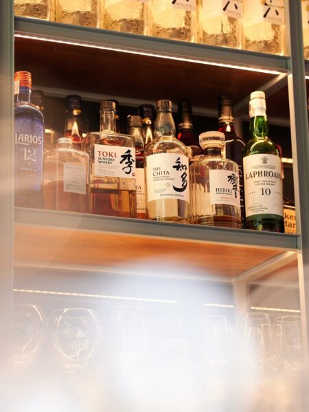 Zbliżenie na półkę z alkoholami w barze – widać kolekcję butelek japońskiej whisky i ginu, m.in. The Chita, Hibiki, Toki oraz Laphroaig. Ciepłe, złote światło oświetlenia akcentuje kolory bursztynowych trunków i nadaje kadr elegancki, luksusowy nastrój.