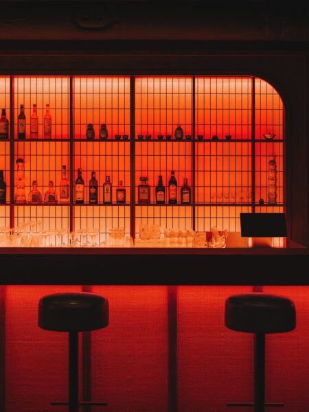 Ciemne wnętrze baru o intensywnym, czerwonym oświetleniu. Na półkach za ladą ustawione butelki alkoholu i szkło, a przed nimi dwa czarne hokery. Światło nadaje przestrzeni futurystyczny, nocny klimat.