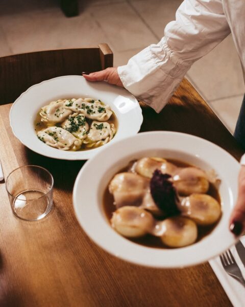 Zbliżenie na dwa talerze z tradycyjnymi daniami kuchni polskiej – pierogi w bulionie z ziołami oraz kluski śląskie z ciemnym sosem i porcją buraczków. Na pierwszym planie ręka w białym mankiecie podaje talerze do stołu.