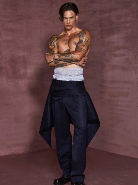 Michał Kot top model 2025: Pozujący, umięśniony model z licznymi tatuażami na torsie i ramionach, ubrany jedynie w ciemne, konstrukcyjne spodnie garniturowe o awangardowym kroju, stojący na tle matowej, winylowej ściany w odcieniu ciemnego różu.