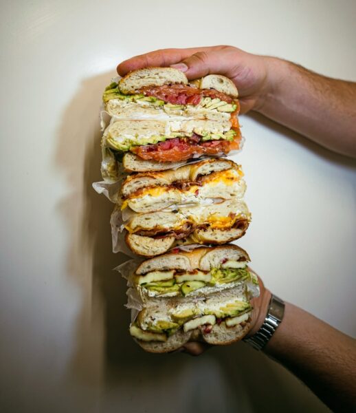 Ujęcie trzech przekrojonych na pół bajgli ułożonych jeden na drugim. Każdy wypełniony różnymi składnikami: warzywami, jajkiem, serem i sosem. Trzymane w dłoniach, pokazują bogactwo struktur i kolorów przekroju.