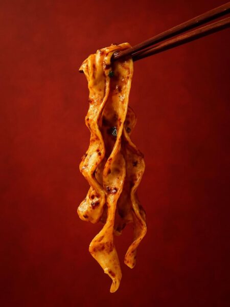 Makrofotografia szerokich, falujących wstążek makaronu pokrytych pikantnym, czerwonym sosem z przyprawami. Danie uniesione pałeczkami na tle w odcieniu ciemnej czerwieni.