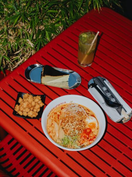 Stół w intensywnie czerwonym kolorze z podanymi daniami: miska pikantnego ramenu z jajkiem, kiełkami i pastą chili, miseczka chrupiących kąsków, kawałek sernika baskijskiego na metalowej tacy oraz wysoka szklanka z mrożonym napojem, w otoczeniu zieleni.