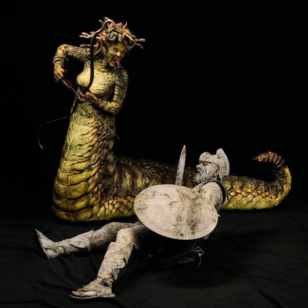 Artystyczna scena inspirowana mitologiczną opowieścią o Meduzie i Perseuszu. Postać Meduzy z długim, gadzim ogonem i łuskowatym ciałem unosi się nad kamiennym wojownikiem z tarczą i mieczem. Kompozycja dynamiczna i dramatyczna, utrzymana w kontrastowym świetle na czarnym tle, podkreślającym fakturę i detale charakteryzacji.