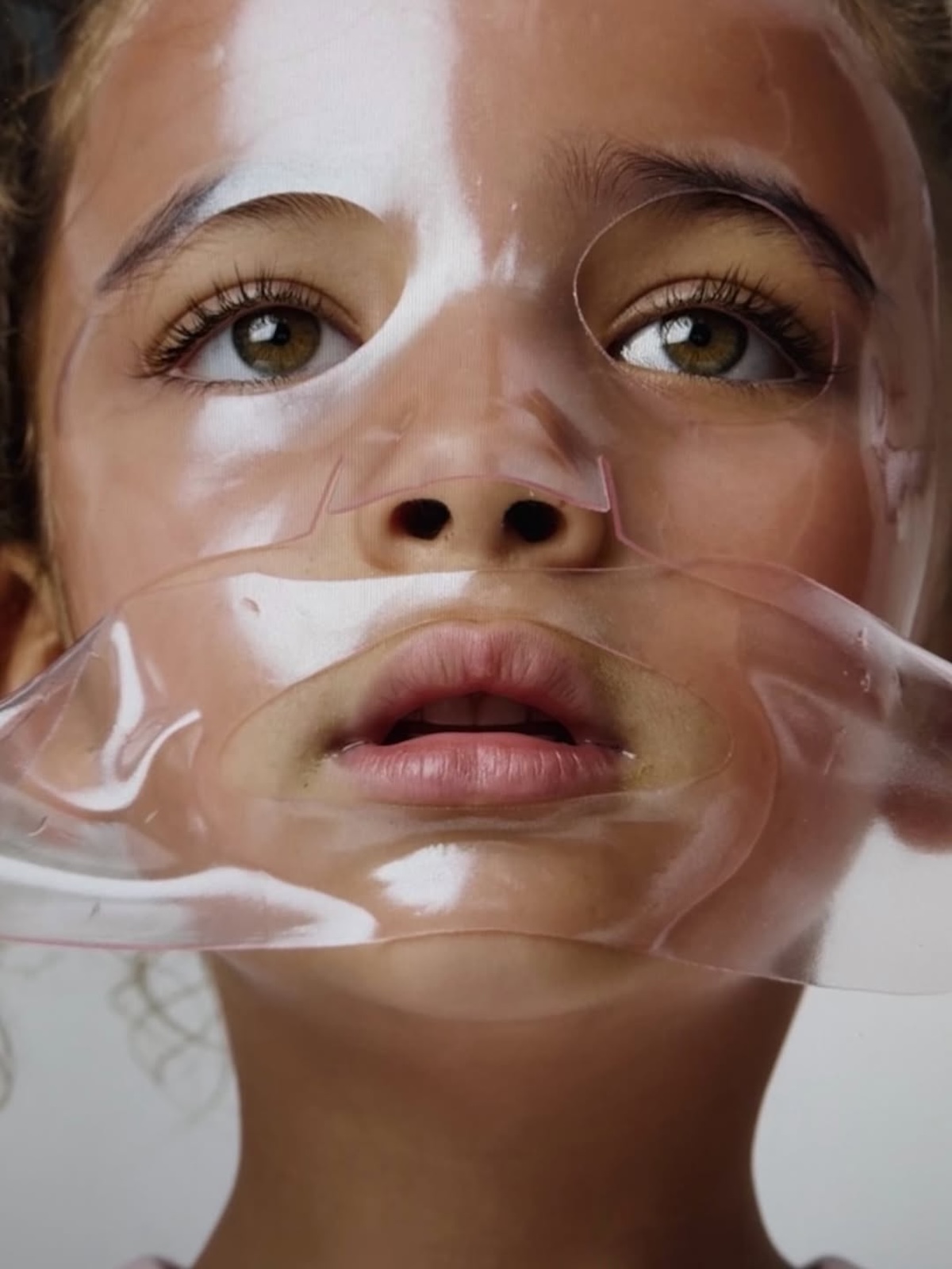Shay Mitchell rini: Zbliżenie twarzy dziecka z nałożoną przezroczystą maską nawilżającą w płacie. Maska dokładnie przylega do skóry, tworząc efekt szklistości i połysku. W kadrze widoczne są duże, brązowe oczy skierowane ku górze oraz naturalna cera o delikatnym blasku – estetyka minimalistyczna, inspirowana trendami pielęgnacyjnymi „glass skin”.