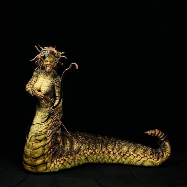 Postać w pełnym kostiumie Meduzy — hybrydy kobiety i węża — przedstawiona na czarnym tle. Łuskowaty ogon zwinięty na ziemi, ciało w odcieniach zieleni, a w dłoniach trzyma długi, zakrzywiony łuk z wężem oplecionym wokół. Ekspresyjny grymas twarzy, wystający język i węże we włosach nadają całości dramatyczny, mitologiczny charakter.