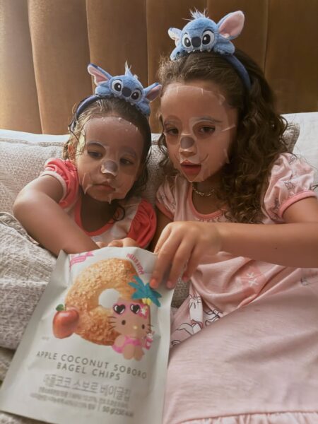 Dwie dziewczynki w piżamach i z opaskami z motywem Stitcha oglądają razem opakowanie przekąsek Hello Kitty. Obie mają na twarzach maski w płacie, a ich skupione spojrzenia i ciepłe oświetlenie tworzą domową, beztroską atmosferę wieczoru spa w dziecięcym wydaniu.