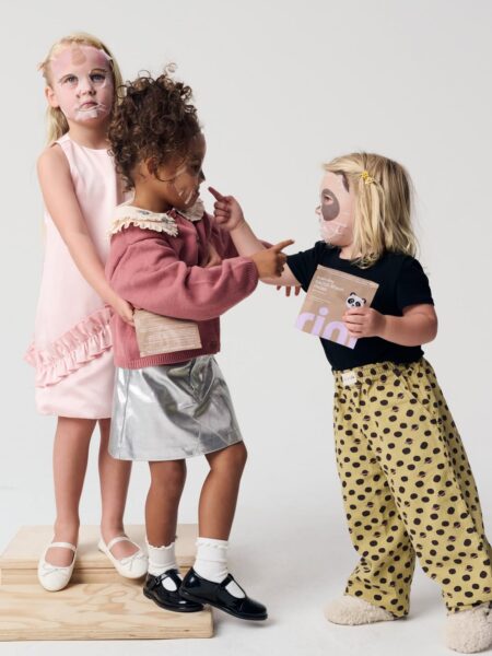 Trzy dziewczynki w maseczkach pielęgnacyjnych stoją razem na jasnym tle studyjnym. Jedna trzyma opakowanie maseczki z wizerunkiem pandy, druga wskazuje na przyjaciółkę, a trzecia obejmuje ją z tyłu. Ich kolorowe stroje — srebrna spódniczka, różowa sukienka i żółte spodnie w grochy — wprowadzają lekkość i radosny ton.